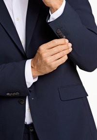 Niebieska marynarka o gładkiej fakturze, z czterema czarnymi guzikami na mankiecie i kieszenią na piersi, w zestawie z białą koszulą elegancką.