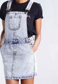 Person som bär en acid-wash denimklänning med framlapp och rullade ärmar på en svart T-shirt, med händerna i sidofickorna.