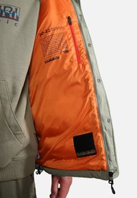 Grüne Puffjacke mit orangefarbener Fütterung; verfügt über eine Reißverschlusstasche, schwarzes Etikett und Druckknöpfe. Der Stoff hat eine glatte Textur.