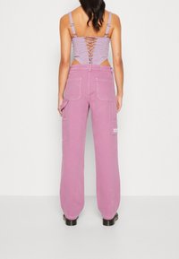 Kvinna iklädd en lila korsettopp med snörning i ryggen och högmidjade rosa cargobyxor, stående mot en enkel vit bakgrund.