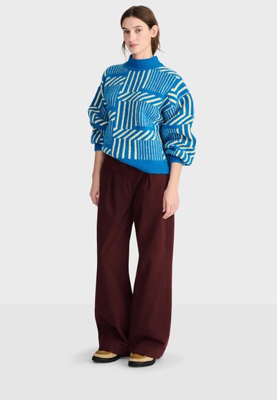 Femme debout portant un pull à motifs bleu et blanc, un pantalon large marron foncé et des chaussures beige sur un fond clair uni.