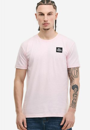 Muž s vrkočmi, oblečený v svetloružovom tričku Lonsdale London a modrých džínsoch, ukazujúci detailné tetovania na pravom predlaktí.