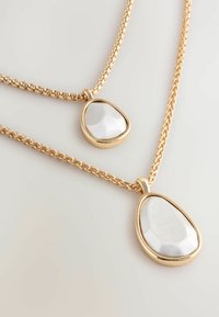 Goudkleurige metalen kettingen met druppelvormige zilveren hangers. Heeft gestructureerde en gladde oppervlakken, met gedraaide schakelverbindingen.