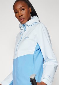 Veste pour femmes en bleu clair et bleu foncé avec une fermeture éclair sur le devant, capuche ajustable et logo sur la poitrine. Texture lisse, design fonctionnel.