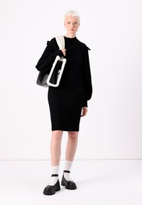 Black ribbed dress with long sleeves, rounded neckline, and shoulder accents. Model carries a black and white fur-trimmed handbag. Chunky black shoes.