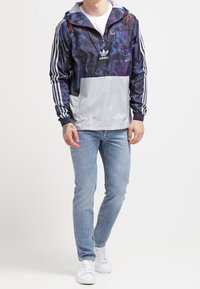 Muž v navy modrém mikině Adidas s galaktickým potiskem a bílými pruhy, světlých modrých džínách a bílých teniskách, stojící s jednou nohou mírně vpřed.