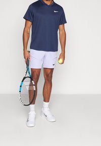 Laivastonsininen urheiluohjelma v-kaula-aukolla, vaalean lila shortsit, joissa on taskut, valkoiset tennarit, pitelee tennismailaa ja palloa.