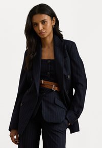 Marineblauwe blazer met krijtstrepen en dubbele knopen, gecombineerd met een bijpassend vest en een bruine riem. Glad stof en gestructureerde silhouet.