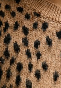 Suéter marrón de punto con una superficie texturizada y manchas irregulares en negro. El cuello es acanalado, lo que resalta el material de punto.