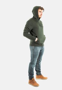 Sudadera verde con un bolsillo frontal, capucha con cordones y puños con rayas blancas, combinada con vaqueros azules y zapatillas altas marrones.