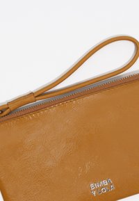 Cartera de piel sintética color marrón claro con una textura suave, cierre de cremallera y una correa para la muñeca. Presenta un logo embellecido en la parte inferior.