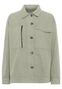Ljusgrön oversizejacka med bruna knappar, en ficka med dragkedja på vänster sida och en klaffficka på höger sida.