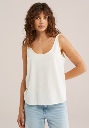 Femme aux cheveux bruns bouclés portant un haut blanc sans manches et un jean bleu clair, debout devant un fond beige uni.