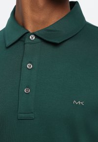 Nærbilde av en person som har på seg en mørkegrønn polo-skjorte med tre sølvknapper og en "MK"-logo brodert på brystet.