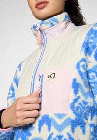 Sudadera de forro polar con un cuerpo acolchado en beige claro y mangas estampadas en azul. Presenta una cremallera rosa y un parche de logo en el pecho.