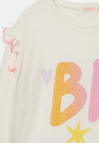 Krémové tričko s glitrovým písmenem "B" v oranžových a růžových odstínech, zdobené růžovým volánkovým lemem na rukávu a malým designem srdce.