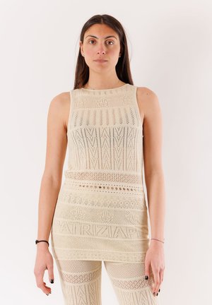 Donna che indossa un abito beige all’uncinetto senza maniche e pantaloni coordinati su uno sfondo bianco.