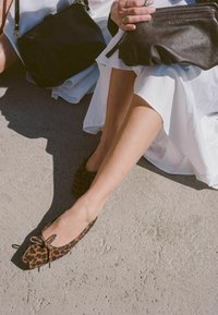 Baleríny s leopardím vzorem a mašlí, vyrobené z jemné látky, s kulatou špičkou, spárované s černými koženými kabelkami, na světle šedém betonu.