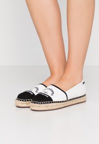 Espadrillas slip-on in tela bianca con punta nera, dettagli neri e branding testuale. La suola in corda di juta aggiunge texture.