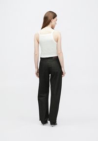 Žena s dlhými hnedými vlasmi, oblečená v bielom tílku, čiernych širokých džínsach a čiernych podpätkoch, stojaca chrbtom k bielej stene.