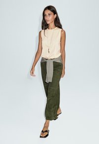 Donna che cammina con un top senza maniche beige, pantaloni verde oliva e un maglione grigio annodato in vita, sandali neri e una lunga collana con pendente.