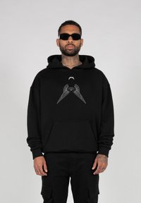 Zwarte hoodie met een zak aan de voorkant, met een zilveren grafisch ontwerp van gestileerde vleugels. Draag het met zwarte cargobroeken en een zonnebril.
