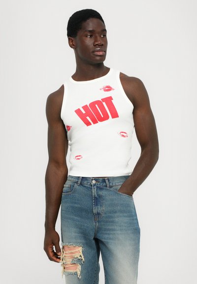 Fehér tanktop piros "HOT" grafikával és csóknyomokkal, amelyhez elhasználódott, kék farmernadrág dukál, szakított részekkel és klasszikus fazonnal.