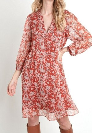 Femme portant une robe orange rouille mi-longue avec des motifs floraux blancs et des manches longues bouffantes, associée à des bottes marron montant jusqu'aux genoux.