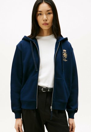 Sudadera con cremallera en color navy y capucha, hecha de tela suave. Presenta un logo dorado en el pecho, bolsillos frontales y puños acanalados.