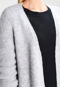 Ljust grått stickat cardigan med öppen framsida, texturerat tyg, ribbade ärmslut, över en svart stickad tröja.