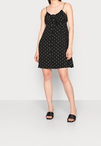 Vestido negro con estampado floral, tirantes finos ajustables, cuerpo fruncido y falda corta. Combinado con sandalias negras de tiras. Textura de tela suave.
