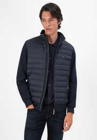 Marineblå puffervest med quiltet tekstur, høy krage og glidelås. Har kontrastfargede ribbestrikkede mansjetter og en liten logo på brystet.