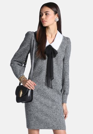 Femme portant une robe en maille grise avec un col blanc et un nœud noir, portant un sac à épaule noir et des bracelets et boucles d'oreilles en or.