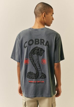 Junger Mann von hinten, trägt ein ausgewaschenes schwarzes T-Shirt mit großem Kobrageschlangengrafik und den Schriftzügen „COBRA“, „Born in the USA“ sowie roten Symbolen.