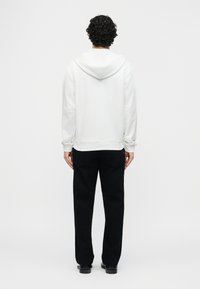 Άτομο με σγουρά μαλλιά που φοράει απλή άσπρη φούτερ με κουκούλα, μαύρο παντελόνι και μαύρα παπούτσια, στέκεται με την πλάτη προς τα εμπρός σε λευκό φόντο.