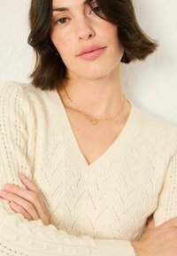 Cremfarbener Pullover mit V-Ausschnitt und strukturiertem Strickmuster. Verfügt über aufwendige Zickzack-Stickerei und gerippte Akzente. Getragen mit einer goldenen Kettenhalskette.