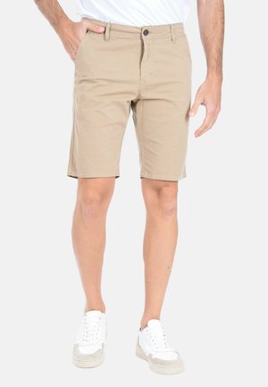 Shorts casual beige indossati con sneakers bianche, mostrati su una persona in piedi con una mano in tasca contro uno sfondo semplice.