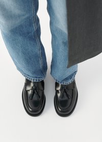 Black leather loafers with tassels, featuring a smooth finish, stitched detailing, and a rounded toe, paired with light blue denim jeans.
