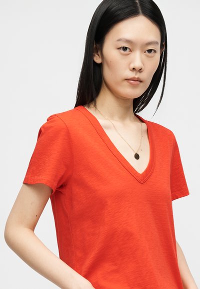 Νέα γυναίκα με ίσια μαύρα μαλλιά που φοράει κόκκινο μπλουζάκι με V-λαιμόκοψη και λεπτή χρυσή αλυσίδα με μικρό στρογγυλό μενταγιόν.