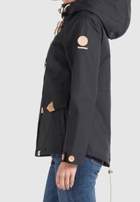 Veste imperméable noire avec capuche, comportant des accents en cuir marron clair, des fermetures à boutons à bascule et des poignets boutonnés. Poches latérales à la forme distinctive.