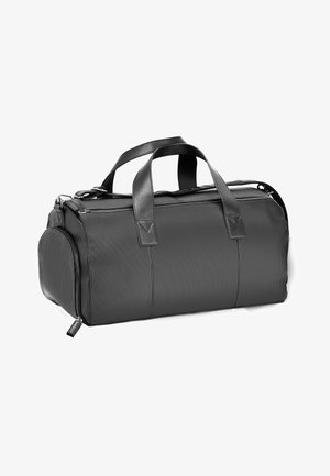 Bolsa de deporte negra con superficie texturizada, doble asa superior y cierre de cremallera. Llevada por una persona con pantalones blancos y zapatillas.