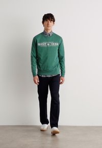 Felpa di cotone verde con testo, indossata sopra una camicia a quadri, abbinata a pantaloni neri e scarpe da ginnastica bianche, in piedi su uno sfondo chiaro.