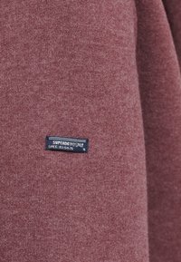 Bordeaux breipapier met een zachte textuur; heeft een blauw label met "SUPERDRY" en specificaties op de stof gestikt.