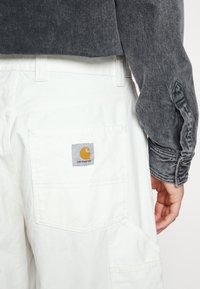 Valkoiset puuvillahaalarit, joissa on suuri tasku takana ja harmaan sekä oranssin Carhartt-logon. Harmaa farkkupaita, jonka hihoissa on tekstuuria.