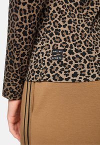 Leopardenmuster-Cordura-Oberteil in Tan und Schwarz, mit einem cropped Design und einem schwarzen Etikett mit der Aufschrift "MARCCAIN SPORTS" an der hinteren Naht.