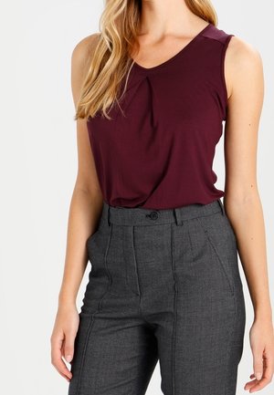Vrouw draagt mouwloze donker bordeauxrode top en hooggetailleerde grijze op maat gemaakte broek met plooien en knoopsluiting.