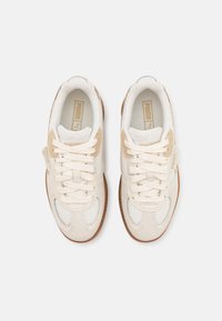 Paire de baskets Puma beiges et blanches avec des détails en mesh et en daim, lacets plats et semelles en caoutchouc marron, vue de dessus sur fond blanc.