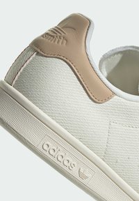 White canvas sneaker featuring a beige leather heel, textured surface, red stitching, and a prominent embossed logo on the side.