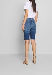 Shorts bermuda de mezclilla en azul oscuro, con dobladillo deshilachado y bolsillos traseros. Combinados con una camiseta blanca y sandalias de tacón marrones.