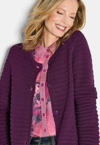 Lila gestrickte Strickjacke mit strukturiertem Design und gerippten Ärmeln, mit runden Knöpfen ausgestattet. Kombiniert mit einer rosa gemusterten Bluse darunter.